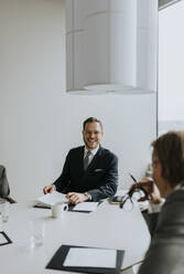 Smiling young businessman looking at colleague while sitting in board room - MASF42342