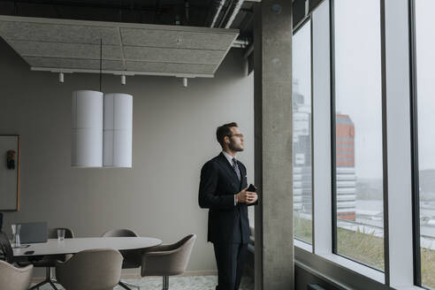 Nachdenklicher Geschäftsmann mit Smartphone, der aus dem Fenster schaut, während er im Sitzungssaal steht - MASF42352