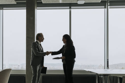 Businessman and businesswoman shaking hands while standing by window at office - MASF42357