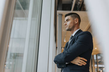 Thoughtful businessman standing with arms crossed looking out through window at office - MASF42391