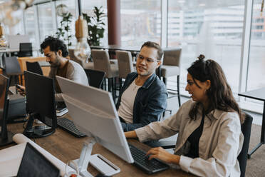 Junge männliche und weibliche Kollegen, die am Computer diskutieren, während sie neben einem Geschäftsmann am Schreibtisch im Büro sitzen - MASF42411