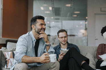 Lächelnder reifer Geschäftsmann mit Kaffeetasse, der während einer Besprechung neben einem männlichen Kollegen sitzt - MASF42425