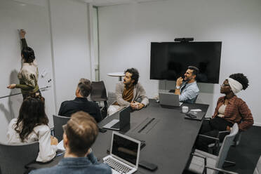 Multirassische Kollegen schauen auf eine Geschäftsfrau, die im Sitzungssaal eine Strategie auf ein Whiteboard schreibt - MASF42445