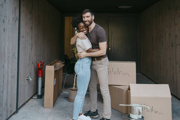 Portrait of happy man embracing girlfriend while standing amidst cardboard boxes - MASF42476