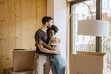 Happy couple embracing each other while standing near window at new home - MASF42482
