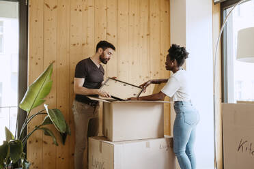 Couple talking to each other while removing frame from cardboard box at new home - MASF42484