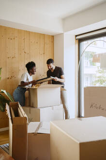 Multiracial couple removing frame from cardboard box while standing at new home - MASF42486