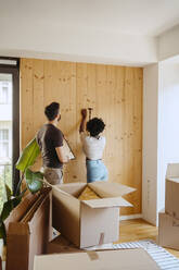 Woman hammering nail on wooden wall while standing with boyfriend at new home - MASF42490