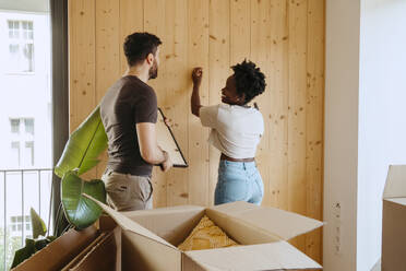 Happy woman hammering nail with boyfriend holding frame near wooden wall at new home - MASF42491