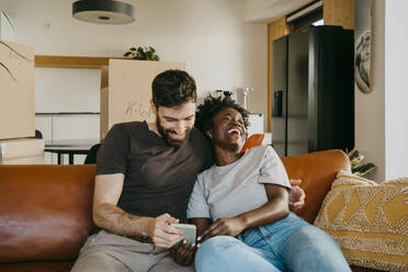 Happy multiracial couple enjoying sharing smart phone while sitting on sofa in living room at home - MASF42498