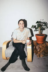 Portrait of smiling woman sitting on chair by plant at home - MASF42537