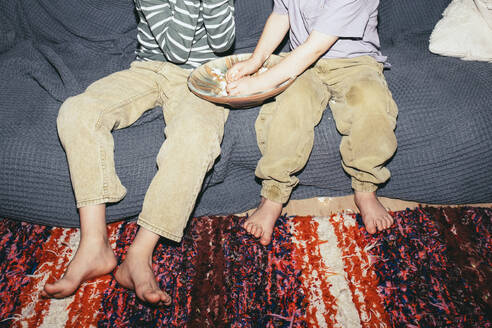 Low section of male siblings sitting with popcorn bowl on sofa at home - MASF42542