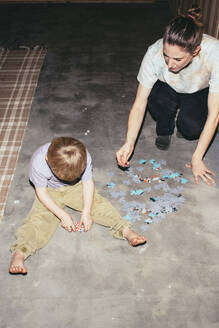Hochformatige Ansicht eines Jungen, der mit seiner Mutter ein Puzzle löst, während er zu Hause auf dem Boden sitzt - MASF42544