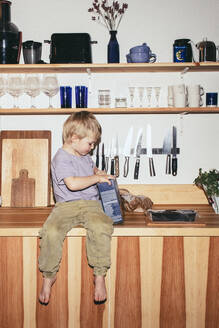 Junge öffnet Lebensmittelpaket, während er zu Hause auf dem Küchentisch sitzt - MASF42545