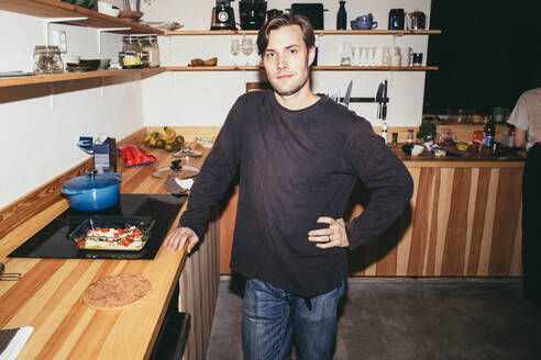 Porträt eines Mannes, der mit der Hand auf der Hüfte steht, während er in der Küche zu Hause Essen zubereitet - MASF42561