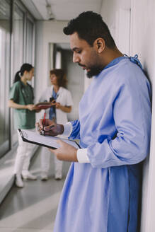 Seitenansicht eines männlichen Chirurgen, der in einem Krankenhauskorridor auf einem Klemmbrett schreibt - MASF42582