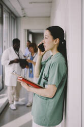 Seitenansicht einer Ärztin, die mit ihrem Smartphone spricht, während sie mit einem Klemmbrett im Krankenhausflur steht - MASF42584