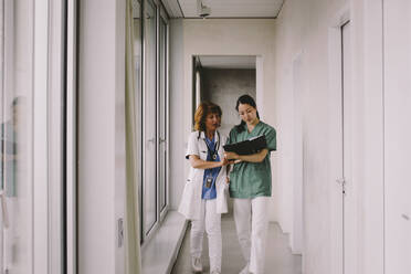 Älterer weiblicher Arzt, der mit einer Krankenschwester über eine Akte diskutiert, während er in einem Krankenhauskorridor geht - MASF42586