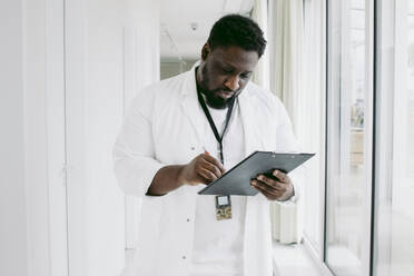 Junger männlicher Arzt schreibt auf einem Klemmbrett, während er im Krankenhausflur steht - MASF42612