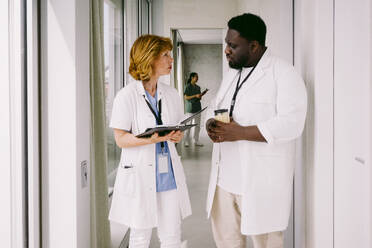 Senior female doctor discussing over medical record with young male colleague while walking in hospital corridor - MASF42616