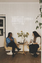 Happy female colleagues discussing while sitting on chairs at office - MASF42641