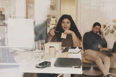 Geschäftsfrau im Gespräch mit einem Kollegen, der im Büro sitzt, gesehen durch ein Glasfenster - MASF42671