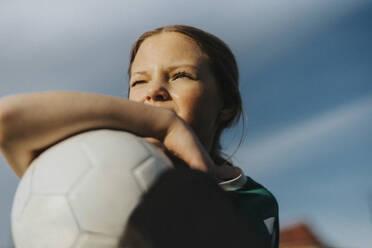 Contemplative elementary girl with sports ball looking away against sky - MASF42693