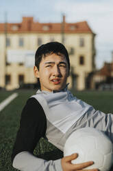 Männlicher Jugendlicher mit Fußball, der auf einem Sportplatz wegschaut - MASF42793