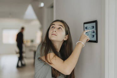 Vorpubertäres Mädchen, das die Raumtemperatur über ein an der Wand befestigtes digitales Tablet zu Hause einstellt - MASF42804