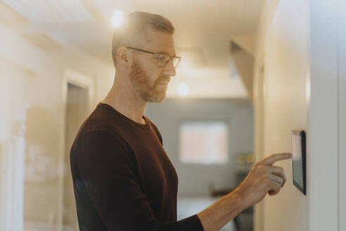 Mature man adjusting room lights through digital tablet mounted on wall at modern home - MASF42810