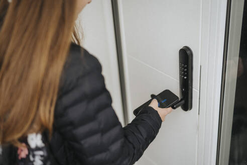Mittelteil eines Mädchens, das zu Hause stehend mit seinem Smartphone das Türschloss entriegelt - MASF42814