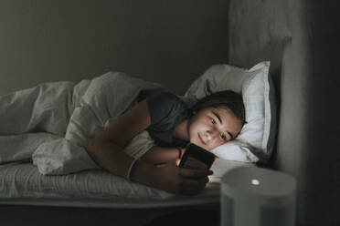 Vorpubertäres Mädchen, das ein Smartphone benutzt, während es im Schlafzimmer auf dem Bett liegt, in einem modernen Haus - MASF42843