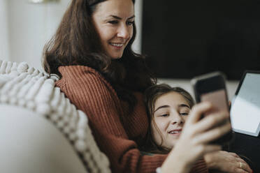 Girl sharing smart phone with smiling mother while lying on sofa in living room - MASF42852