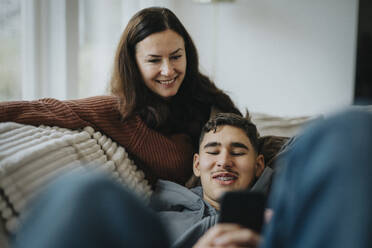 Smiling boy sharing smart phone with mother while lying on sofa in living room at home - MASF42853