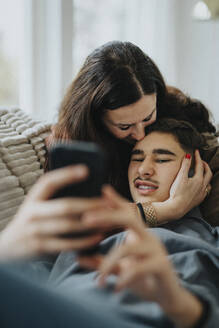 Teenager, der ein Mobiltelefon benutzt, während seine Mutter ihn im Wohnzimmer küsst - MASF42854