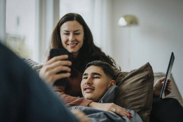Teenage boy sharing smart phone with mother while lying on sofa in living room at home - MASF42855