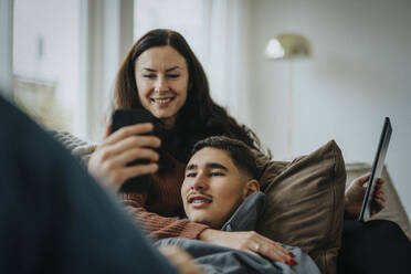 Teenager, der sein Smartphone mit seiner Mutter teilt, während er zu Hause auf dem Sofa im Wohnzimmer liegt - MASF42855