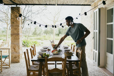 Junger Mann deckt den Esstisch für eine Dinnerparty auf der Terrasse - MASF42891