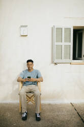 Voller Länge des jungen Mannes mit Smartphone sitzt auf Stuhl gegen weißes Haus - MASF42894