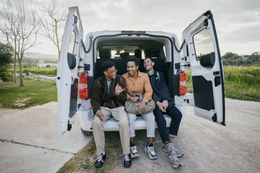 Full length of cheerful male friends sitting in van trunk on roadside - MASF42905