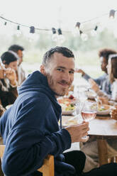 Porträt eines lächelnden reifen Mannes, der mit einem Weinglas auf einer Dinnerparty im Innenhof sitzt - MASF42933