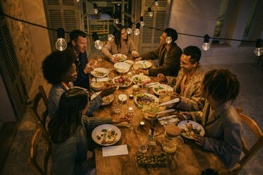 Hoher Blickwinkel auf lächelnde Freunde, die an einem beleuchteten Esstisch während einer Dinnerparty sitzen - MASF42946