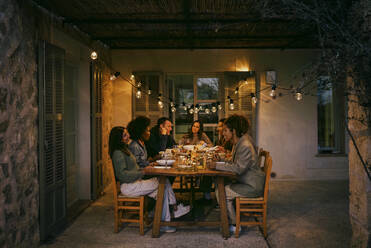 Multirassische männliche und weibliche Freunde sitzen am Esstisch im Innenhof während einer Dinnerparty - MASF42997