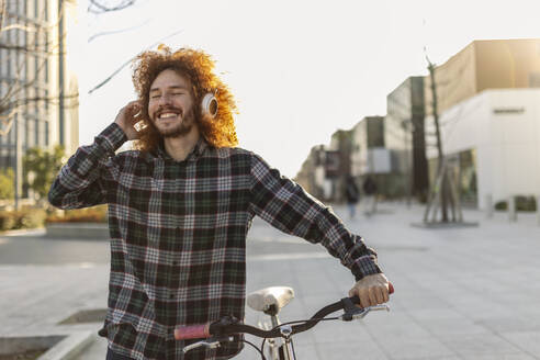 Happy man listening music through wireless headphones and walking with bicycle - JCCMF11111