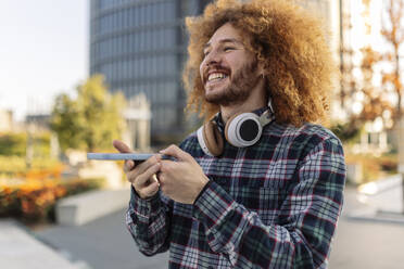 Happy curly haired man with smart phone and wireless headphones - JCCMF11114