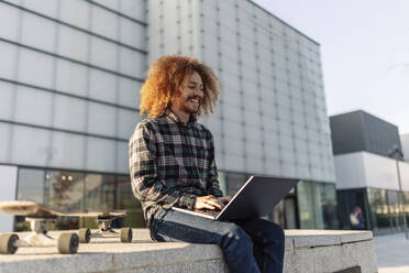 Happy freelancer working on laptop in front of building - JCCMF11117