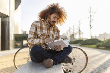 Man reading book and sitting on seat near bicycle - JCCMF11120