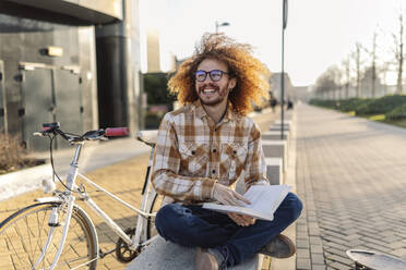 Smiling man with book sitting on seat near bicycle - JCCMF11121