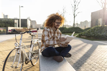 Happy man reading book on seat near bicycle - JCCMF11122