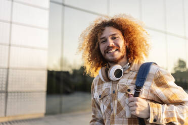 Happy redhead man with curly hair near building - JCCMF11147